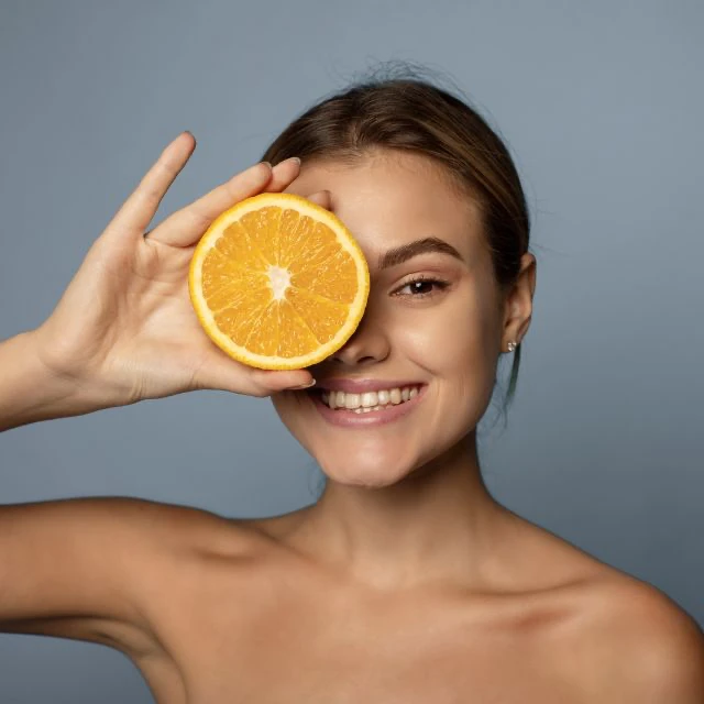 Joven sonriente sujetando media naranja delante de su ojo derecho